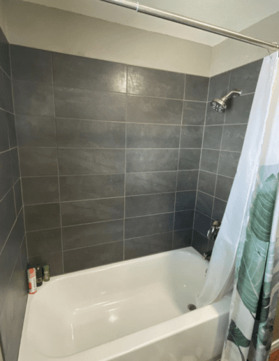 A white bathtub with a showerhead and gray tiled walls. There is a patterned shower curtain partially drawn, and a few shampoo bottles are placed in the corner of the tub.