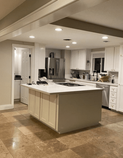 A modern kitchen with white cabinets, a central island, stainless steel refrigerator, and tan tiled flooring. The kitchen has recessed lighting and a door leading to another room.