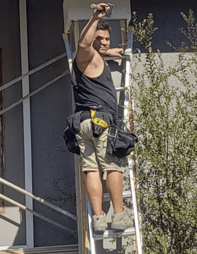 A person wearing a black tank top and beige shorts works on a ladder, painting a building's exterior trim.