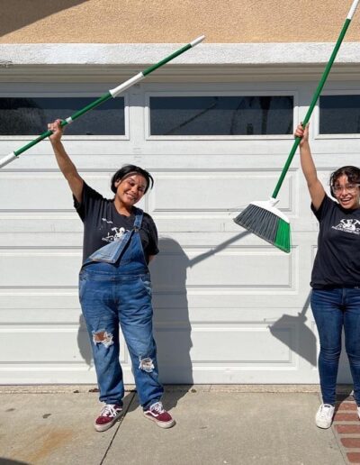 Two people stand in front of a garage door, smiling and raising a mop and a broom. They are both wearing black t-shirts and jeans.