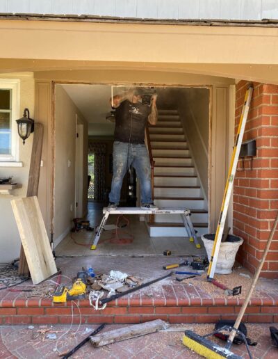 A person standing on a stepladder uses a power tool to work on the upper frame of a doorway under construction with various tools and materials scattered on the ground.