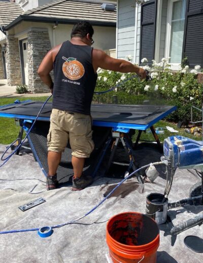 A man wearing a black tank top and khaki shorts is spray painting a large panel supported by sawhorses outdoors. A paint machine, bucket, and other painting equipment are on the ground nearby.