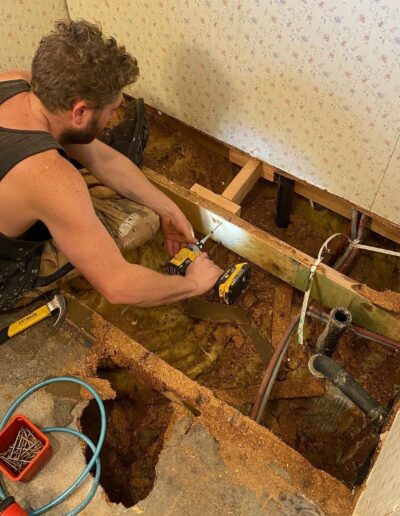 A person wearing a black sleeveless shirt uses a power drill to fix wooden support beams in a partially demolished section of a room, with tools and materials scattered around.