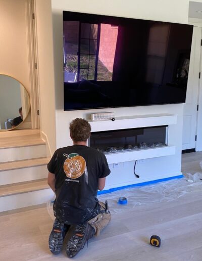 A person kneels on the floor working on the wiring below a mounted flat-screen TV in a living room. Tools and a protective sheet are spread out around them.