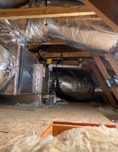 An attic space with exposed wooden beams, insulation, and various HVAC ductwork and pipes. Some insulation is laid on the floor, and a section of the floor panel is missing.