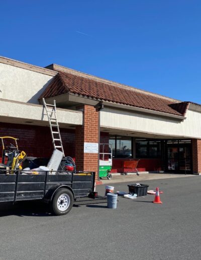 A worker on a ladder repairs the exterior of a building with a "BIG" sign, while tools and equipment are scattered in a trailer and on the ground in front of the store.
