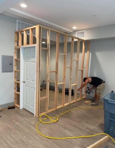 A person is working on constructing a wooden framework around a door inside a room. Various construction materials and tools are scattered around, with an extension cord on the floor.