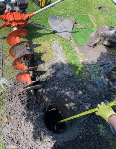 A person wearing a yellow glove measures the diameter of a hole in the ground using a tape measure. An earth auger used to dig the hole is visible on the grass beside it.
