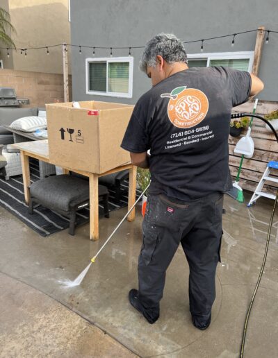 A man pressure washes a concrete surface near a table with a large cardboard box on it in a backyard, wearing a black t-shirt and dark pants.