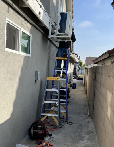 A person stands on a ladder working on an air conditioning unit mounted on the side of a building. Various tools and equipment are scattered on the ground below.