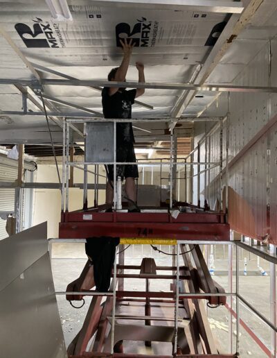 A person stands on a scissor lift, installing insulation panels on a ceiling in a partially constructed building.