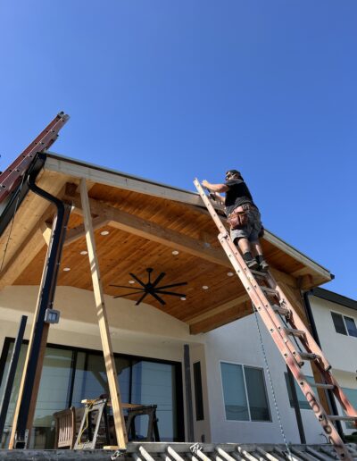 A man is standing on a ladder and working on the wooden ceiling of a covered outdoor patio. The sky is clear and blue. There are two ladders and construction materials in the scene.