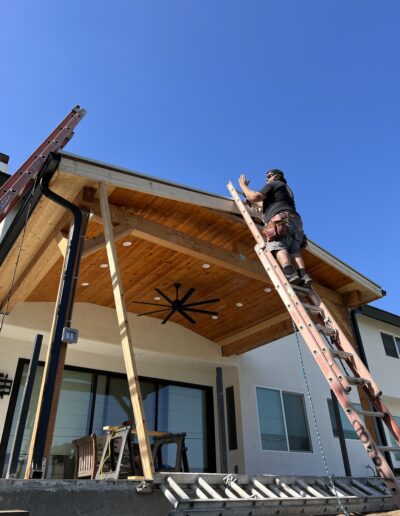 A person stands on a ladder working on the roof of a house with wooden patio ceiling under a clear blue sky. Construction materials are visible on the patio below.