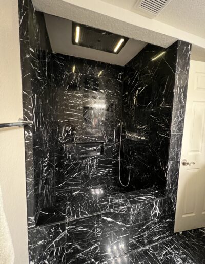 A luxurious bathroom features a black marble shower area with white veining, overhead rain shower, handheld showerhead, and wall-mounted shower controls.
