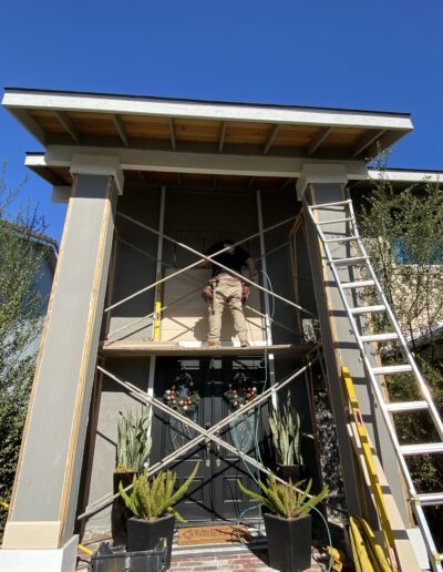 A person is standing on scaffolding while doing construction work on a two-story house's exterior entryway, with a ladder leaning on the side and potted plants by the front door.