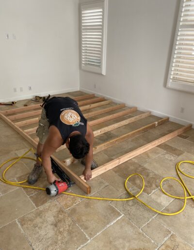 A person wearing a black tank top and khaki shorts is using a nail gun to assemble a wooden frame on the floor of a room with beige tiled flooring and white walls.