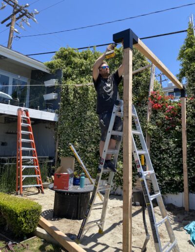 A person stands on a ladder working on a wooden structure in a backyard. Multiple ladders, tools, and a cardboard box are seen nearby.