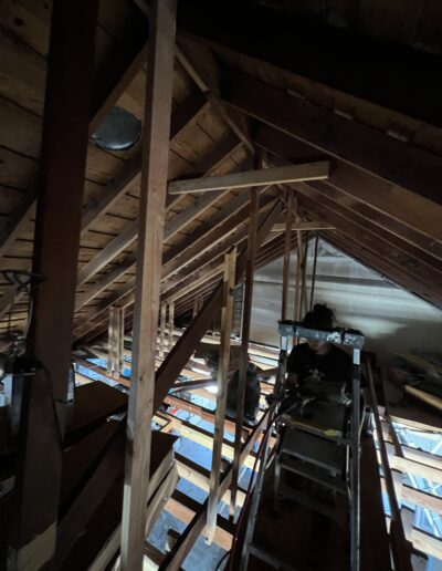 A person is working in an attic, standing on a ladder among exposed wooden beams and various stored items. The lighting is dim.