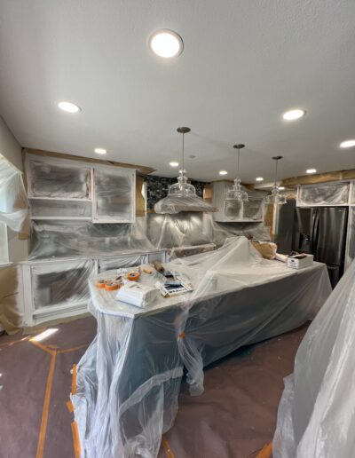 A kitchen undergoing renovation, with plastic sheets covering the cabinets, countertops, and appliances. Various tools and materials are scattered on the countertop and floor.