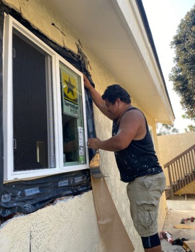 A person in a black tank top and khaki shorts is installing a window on the exterior of a beige building. A brown paper is held up for guidance, and various tools and materials are visible.