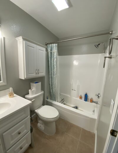 A clean bathroom featuring a white vanity, toilet, and bathtub with a shower curtain. The walls are painted light gray, and the floor is tiled. Various toiletries are visible on the bathtub ledge.