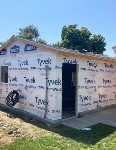 A small house under construction with Tyvek HomeWrap on the exterior, a coil of tubing on the ground, and nearby tools. A wooden fence and trees are in the background.