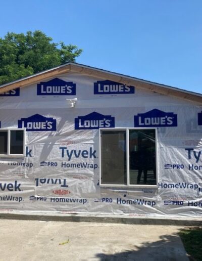 A small house under construction with Tyvek HomeWrap and Lowe's branding on the exterior. A window and a glass sliding door are installed. A ladder is positioned on the left side.