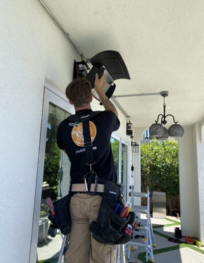 A technician in a tool belt installs an outdoor light fixture under a covered patio. A ladder and various tools are visible in the background.