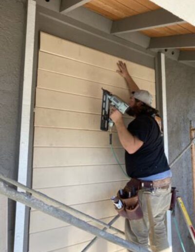 Person wearing a tool belt and hat installs wooden siding on an exterior wall using a nail gun. Scaffolding and construction tools are visible around them.