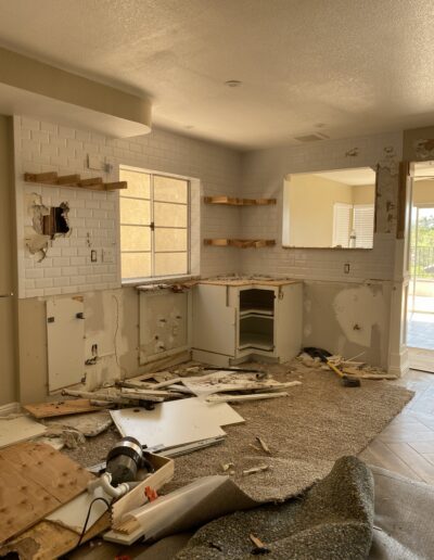 A kitchen under renovation with cabinets removed, tools scattered, open wall spaces, and exposed plumbing. A large mirror and window are visible, and debris is spread on the floor.