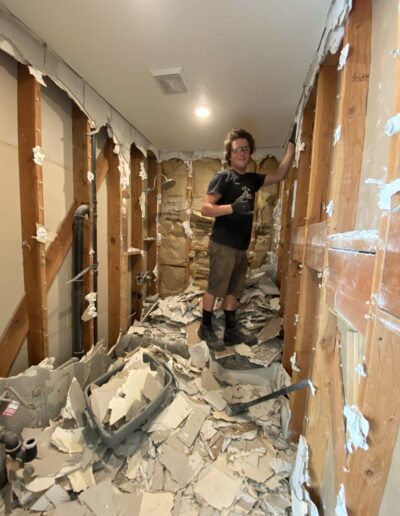 A person stands amidst debris in a stripped-down room, with exposed wooden studs and demolition material scattered across the floor.