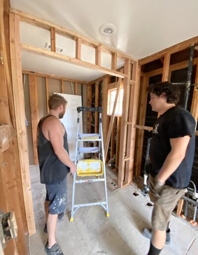 Two men working on a home renovation project, standing in a room with exposed wooden framing and a yellow ladder. One man is gesturing towards the ceiling.