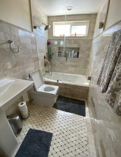 A bathroom with a toilet, sink, and bathtub-shower combination. The space has beige tiled walls and floor, a glass shower door, a towel holder with towels, and two dark mats on the floor.