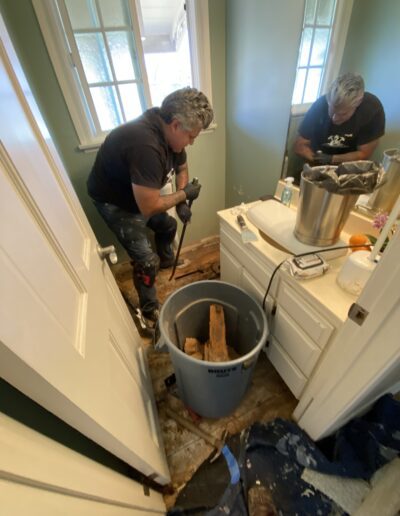 A person working on bathroom floor repairs, removing damaged wood, with a trash can and tools nearby.