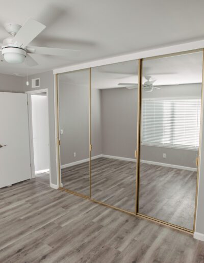 A bedroom with gray walls, a ceiling fan, mirrored sliding closet doors, wood-like flooring, two doors (one open and one closed), and a window with horizontal blinds.