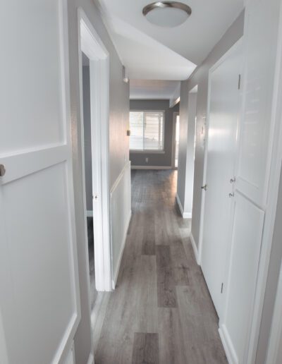 A narrow, well-lit hallway with light gray walls, white doors, and a gray wood floor leading to a room with a window.
