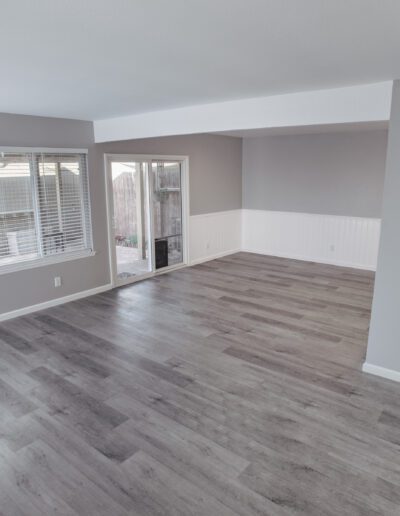 A spacious empty room with light grey walls, large windows, sliding glass door, and dark wood floors.