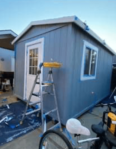 A small gray shed with a white door and window is being painted. A ladder, paint supplies, and a bicycle are in front of the shed.