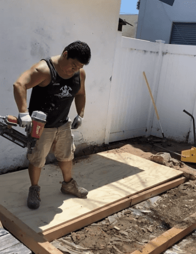 Person in a black tank top and khaki shorts using a nail gun to build a wooden structure in a backyard with white fencing.
