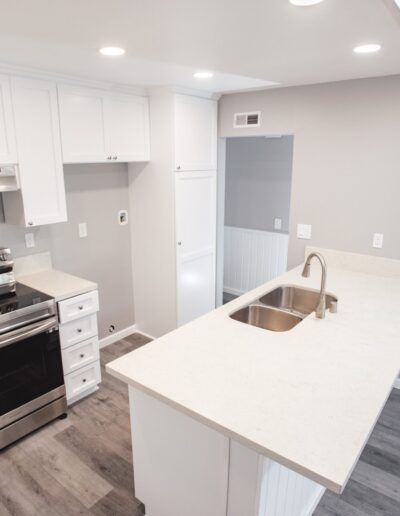Modern kitchen with white cabinets, stainless steel appliances, and a large island with a sink. The space features gray walls and wood flooring, with recessed lighting illuminating the area.