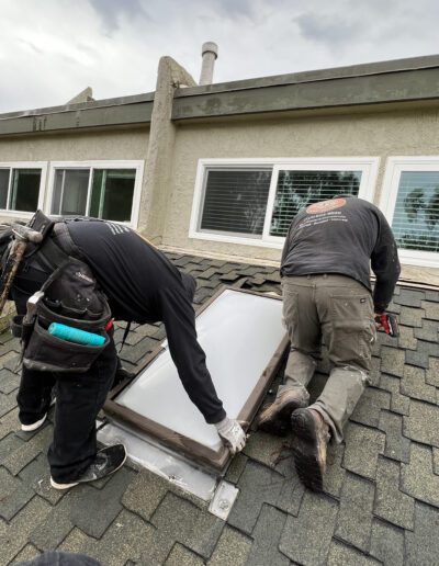 Two workers in dark clothing kneel on a roof while installing a skylight. One handles the frame, and the other, holding a tool, works on securing it in place.