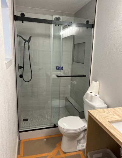 A modern bathroom with a glass-enclosed shower, white toilet, toilet paper rolls on the tank, and a partially unfinished wooden vanity.