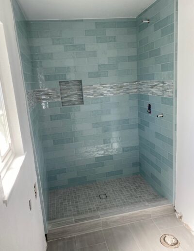 A newly tiled shower with blue-green subway tiles, a mosaic accent strip, and inset shelf. The floor has small square tiles, and the rest of the bathroom floor is tiled in a light gray color.
