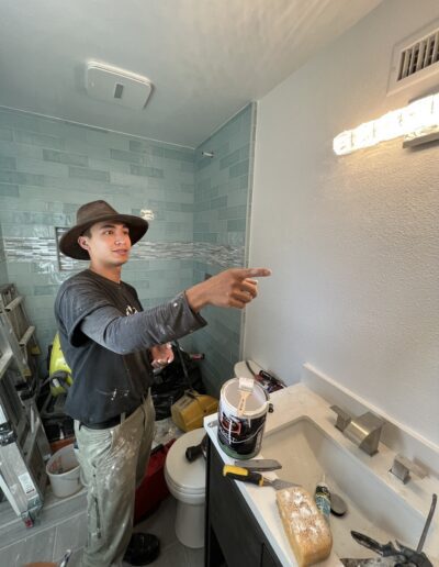 A person wearing a hat and work clothes is pointing at something on the wall of a bathroom under renovation. A ladder, tools, and paint supplies are visible.