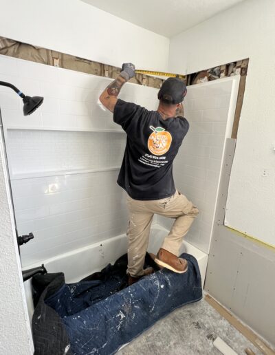 A worker wearing gloves and a cap measures tiles above a bathtub in a bathroom under renovation, with a blue tarp covering part of the tub.