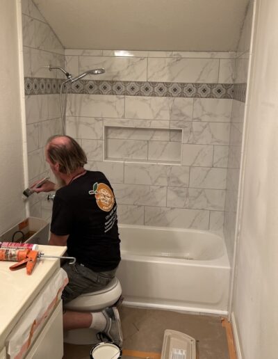 A person works on tiling a bathroom wall above a bathtub, wearing a black shirt and shorts, with tools and paint supplies around. A step ladder and partially opened cabinet are visible.