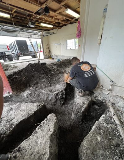 A worker kneels beside a large excavation hole inside a garage. The hole appears to be under construction or repair with tools and soil scattered around the area.
