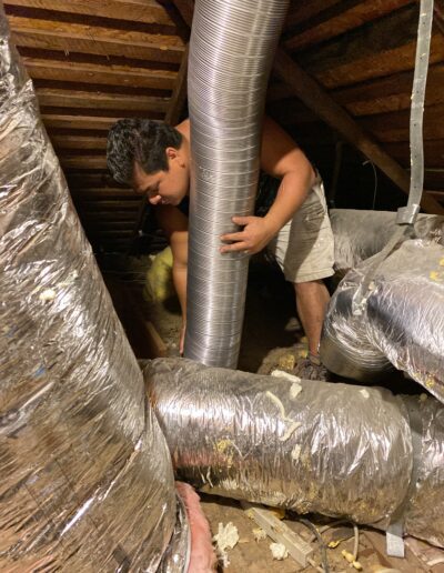 A person adjusts a large, flexible duct in an attic space surrounded by various other air ducts and insulation.
