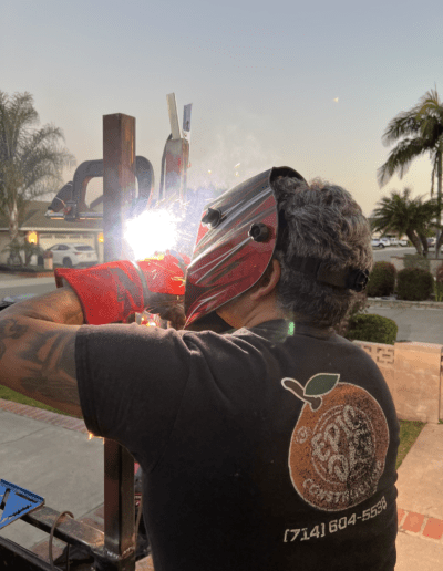 A person wearing a welding mask and gloves welds a metal structure outside. Palm trees and a suburban street are visible in the background.