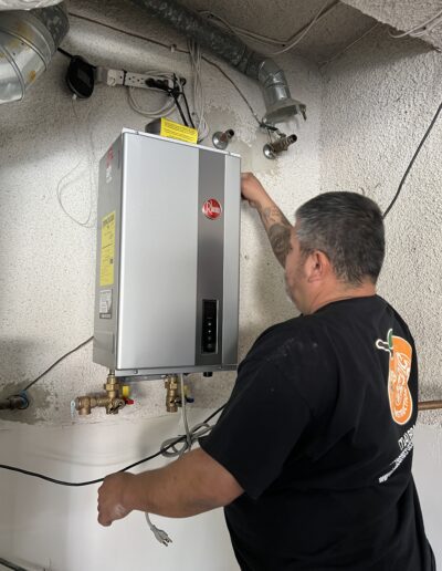 A person with tattoos wearing a black shirt is adjusting a water heater mounted on a wall in a utility room with visible pipes and wires.